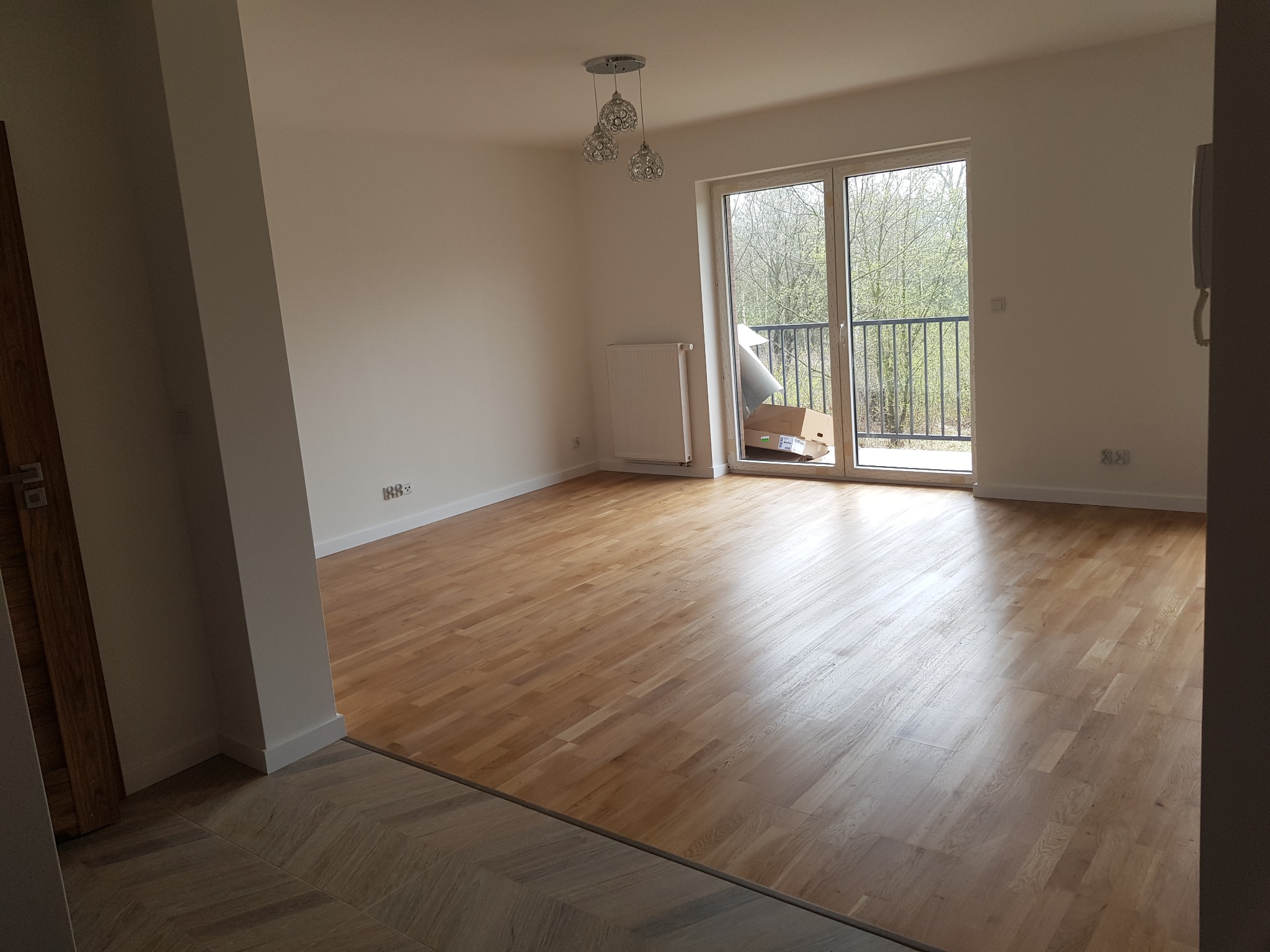 Jasne, puste pomieszczenie z nową, świeżo ułożoną drewnianą podłogą w jodełkę przechodzącą w klasyczny parkiet. Widoczne okno balkonowe z widokiem na drzewa i grzejnik na ścianie. Na balkonie kartony.