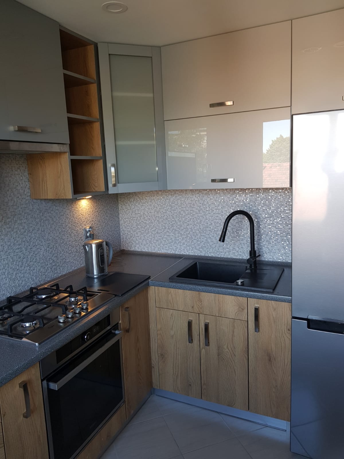 Nowoczesna kuchnia z drewnianymi szafkami, czarnym zlewem i armaturą, oraz mozaikową ścianą nad blatem, oświetlona punktowym światłem