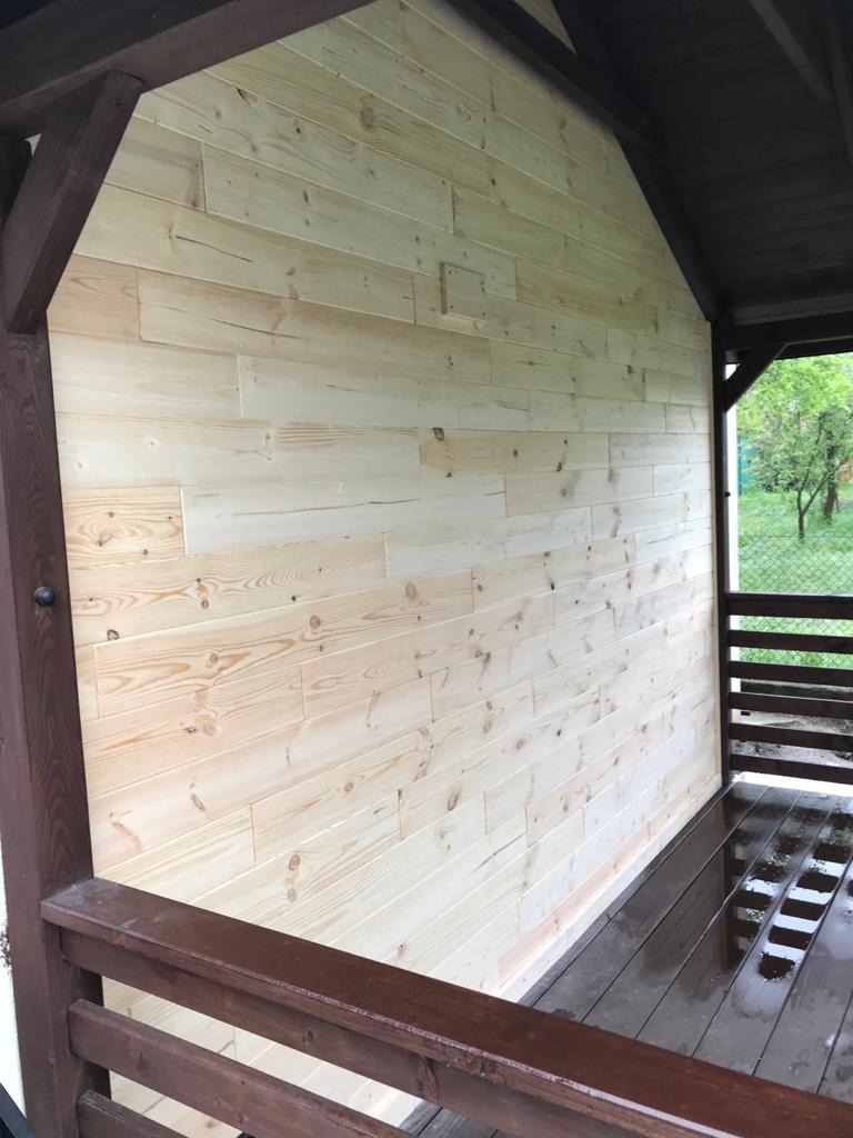 Ściana altany wykończona jasnymi panelami drewnianymi, widok z zewnątrz na mokry drewniany podest i zieleń w tle.