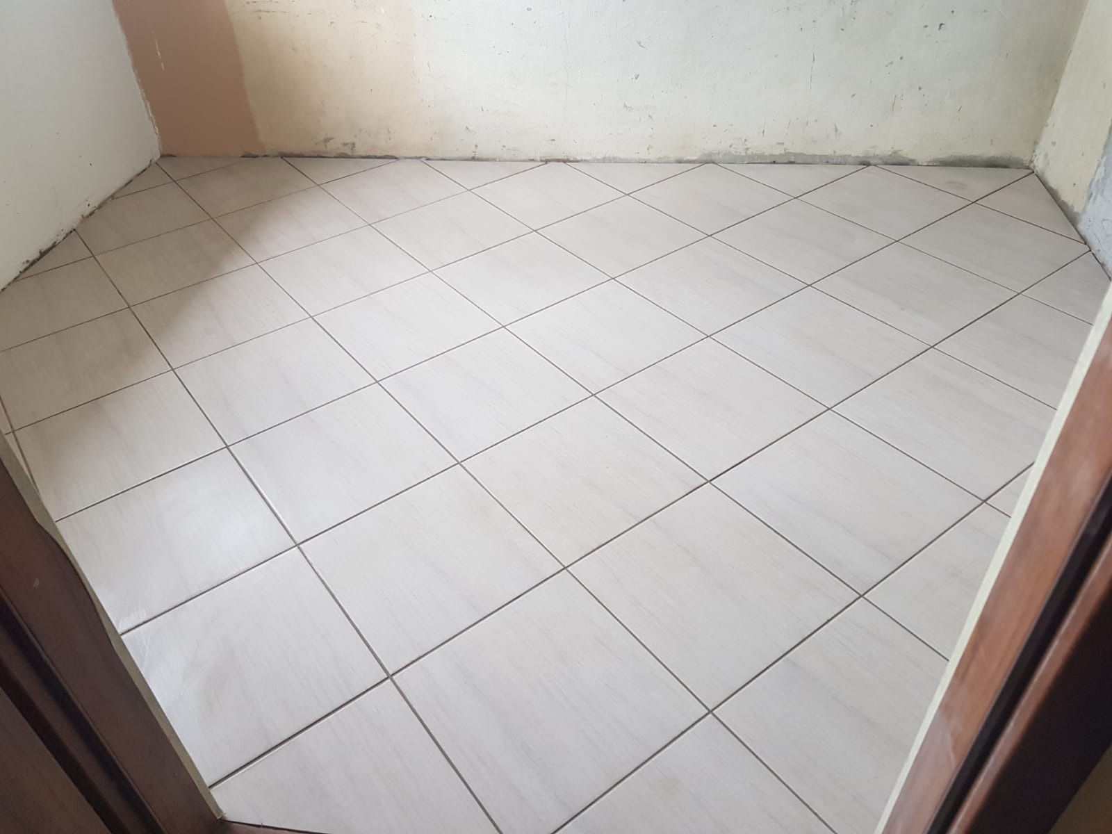 Jasne płytki podłogowe ułożone ukośnie w małym pomieszczeniu, widoczne surowe ściany w trakcie remontu i fragment drewnianej ościeżnicy drzwi.