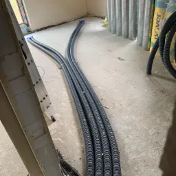Szare, elastyczne rury wentylacyjne ułożone na surowym betonowym podłożu w trakcie instalacji, widoczne również rury spiro i drabina oparta o ścianę.