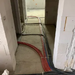 Wnętrze w trakcie budowy z widocznymi przewodami instalacji grzewczej i hydraulicznej, prowadzonymi po podłodze i mocowanymi do ścian z betonu komórkowego.