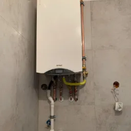 Biały piec gazowy Termet zamontowany na ścianie z szarych płytek, widoczne miedziane rury, żółty przewód gazowy oraz białe rury odpływowe, obok gniazdko elektryczne.