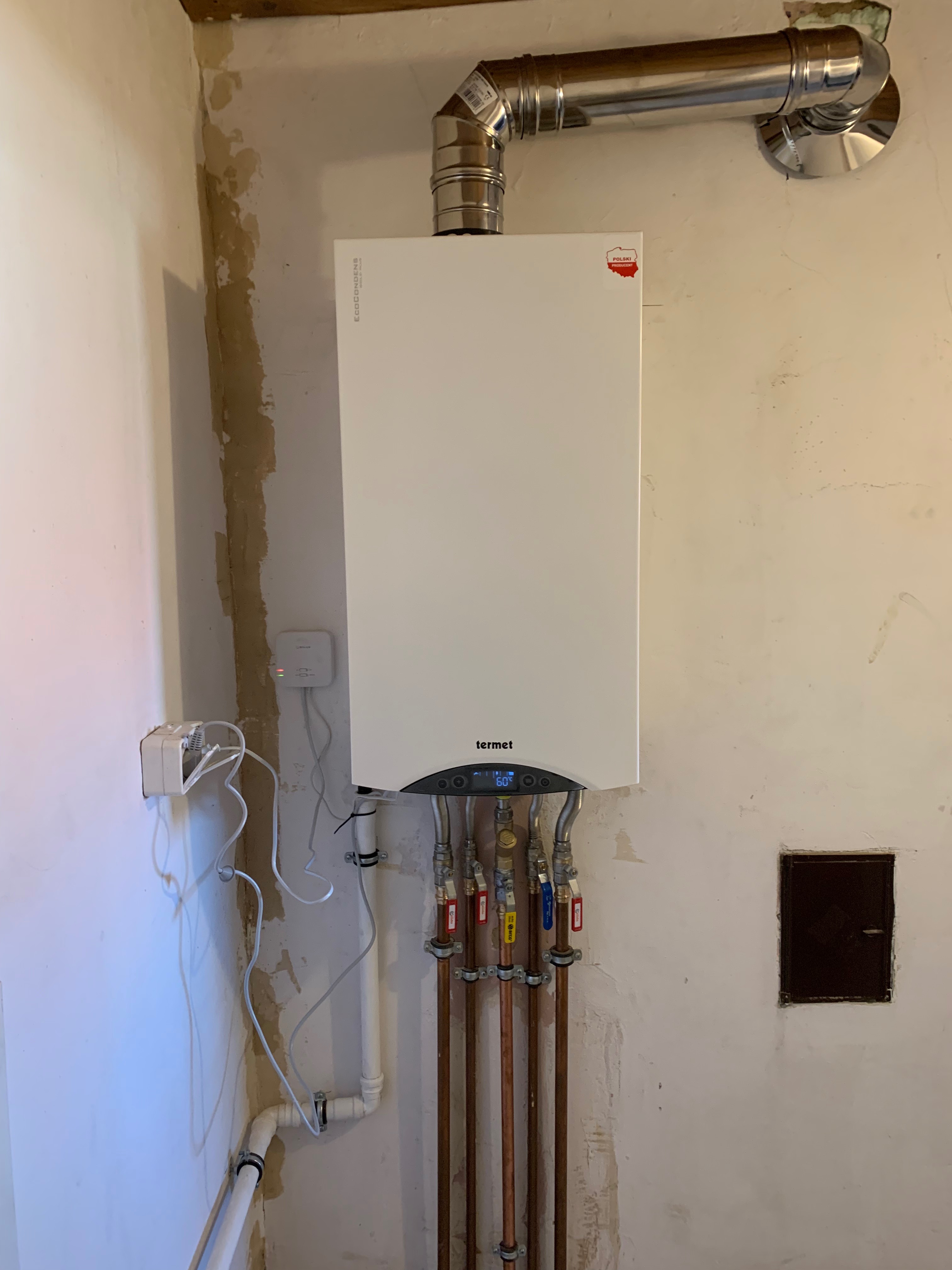 Nowy, biały piec gazowy marki Termet zamontowany na ścianie w trakcie remontu. Widoczne rury miedziane z zaworami, przewody odprowadzające spaliny oraz niedokończone wykończenie ściany.