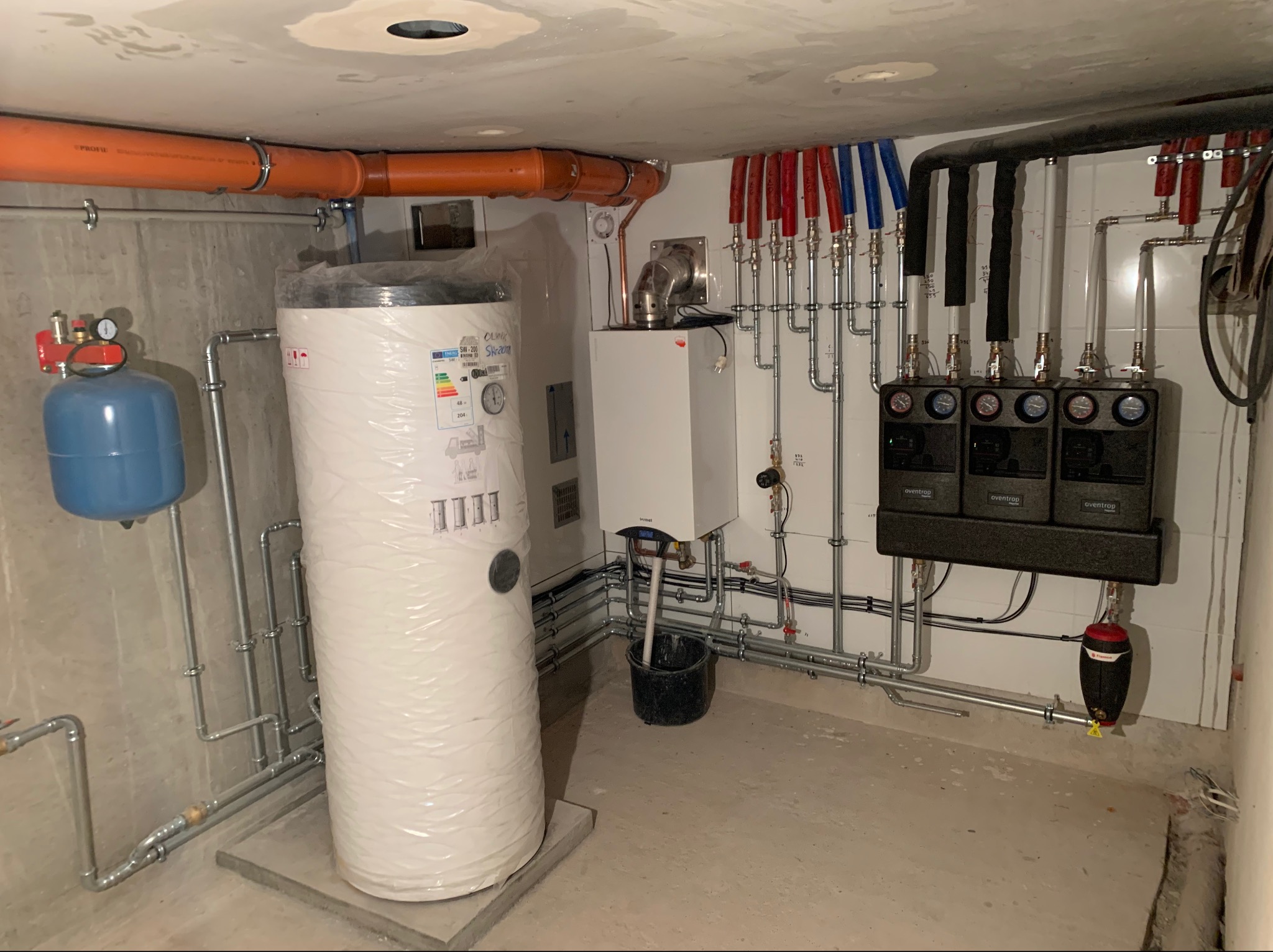 Kotłownia w trakcie montażu: biały, owinięty folią zasobnik ciepłej wody na betonowym podeście, obok szary piec, orurowanie w ścianach, trzy czarne moduły z manometrami Oventrop, ekspansomat w tle,...