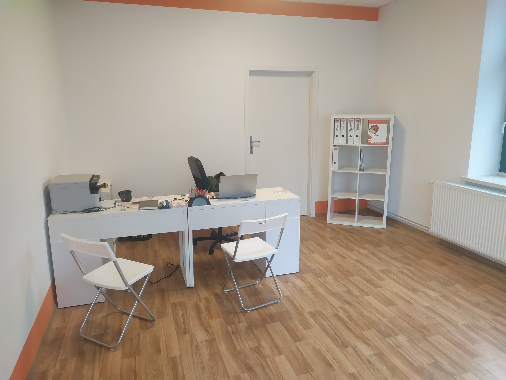 Jasne biuro z białymi meblami: biurko z laptopem, drukarką i krzesłami, regał z segregatorami, drewniana podłoga, pomarańczowy akcent kolorystyczny na suficie.