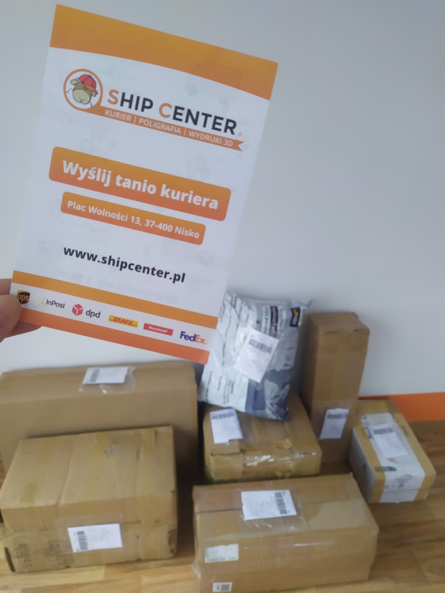 Ulotka firmy kurierskiej Ship Center z adresem i logiem, trzymana w dłoni, na tle paczek kurierskich różnych firm.