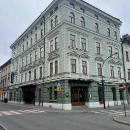 Odrestaurowana kamienica narożna w odcieniach zieleni z białymi zdobieniami, widoczna z perspektywy ulicy z przejściem dla pieszych.