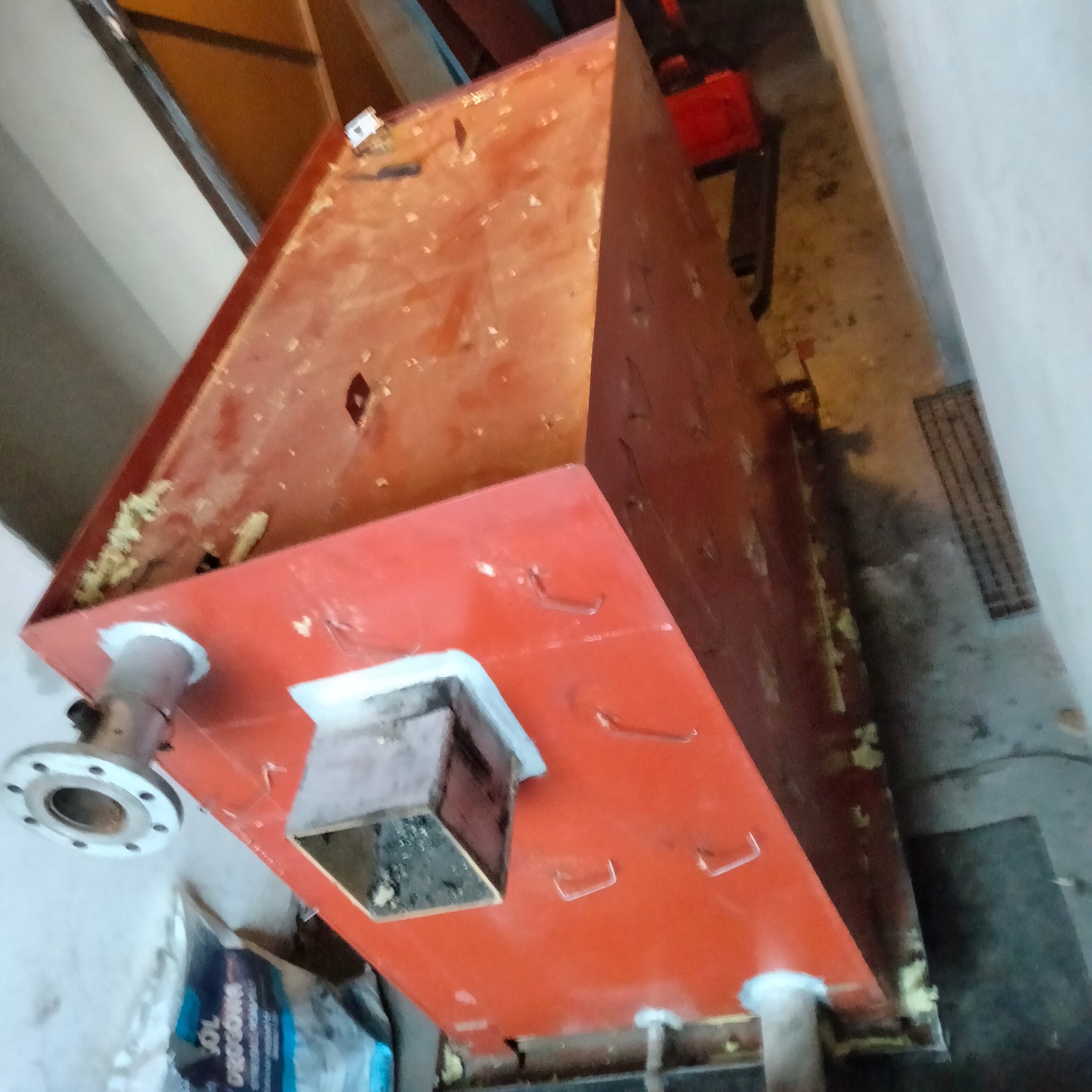 Czerwony, metalowy zbiornik o nietypowym kształcie, z widocznymi elementami instalacji i śladami użytkowania, ustawiony w pomieszczeniu gospodarczym.