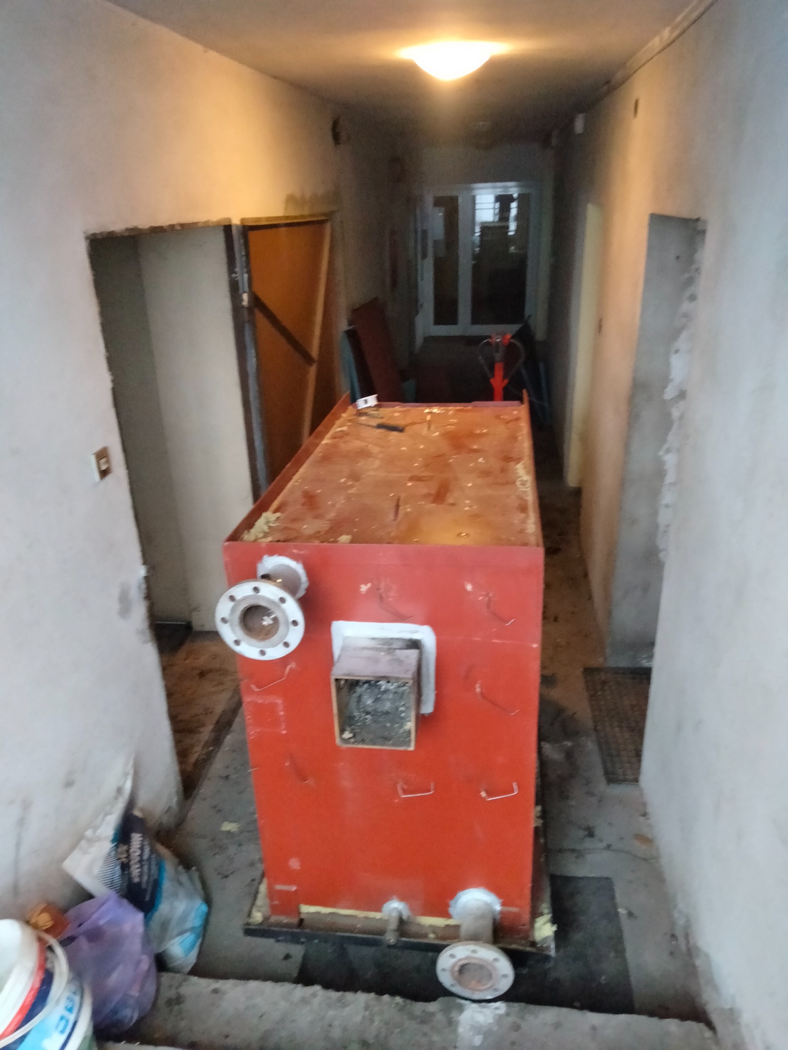 Czerwony piec centralnego ogrzewania z przyłączami, umieszczony w wąskim korytarzu w trakcie demontażu, widoczne worki z resztkami materiałów budowlanych i wózek transportowy w tle.