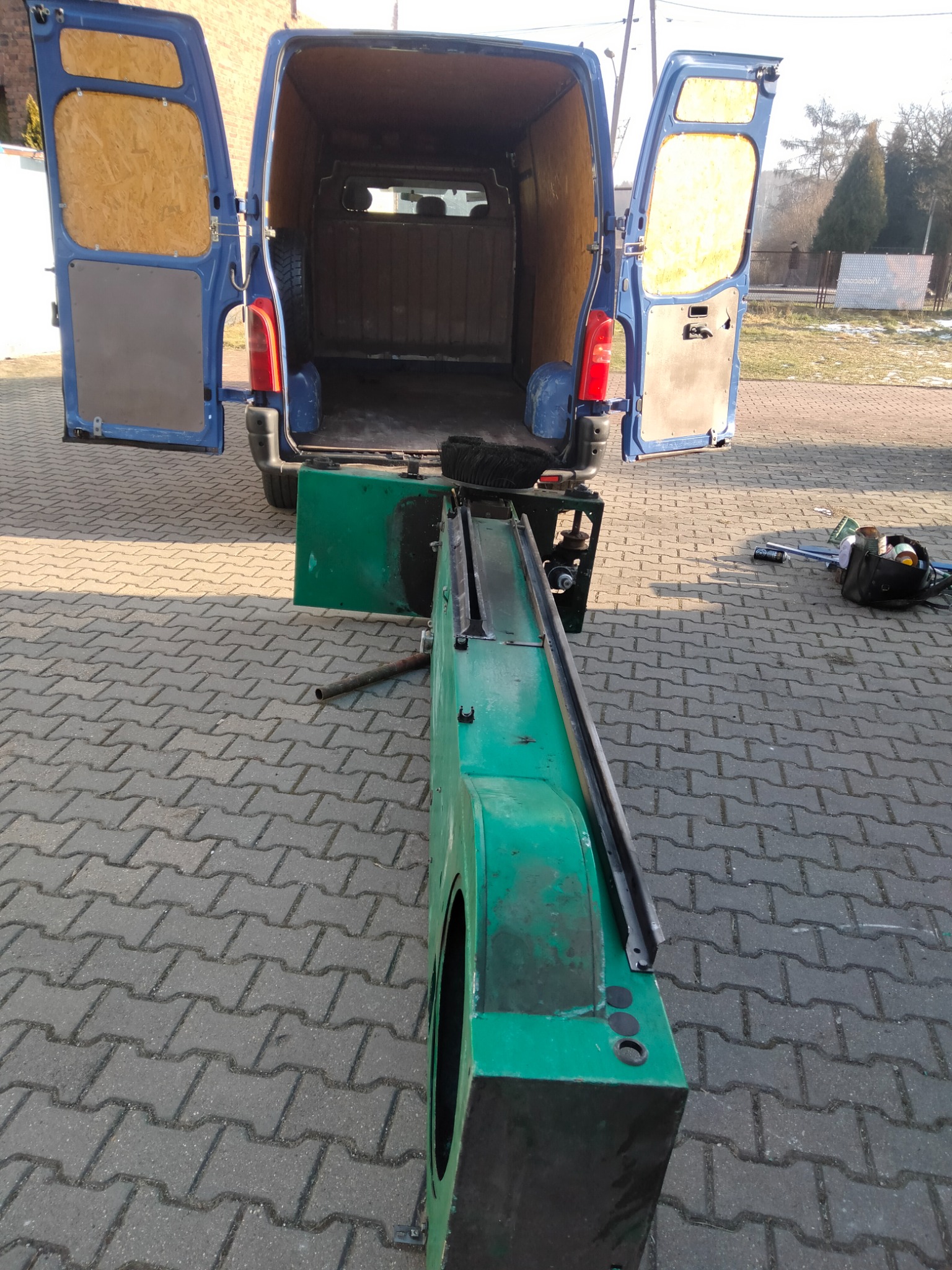 Niebieski bus z otwartymi tylnymi drzwiami, obok zielona konstrukcja z metalowymi elementami i opona wewnątrz pojazdu, na zewnątrz kostka brukowa i torba z narzędziami.