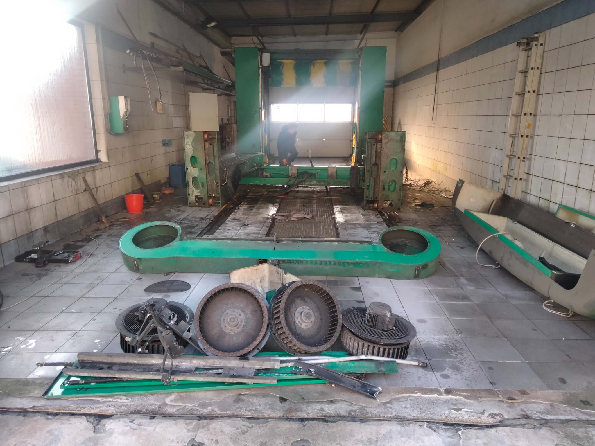 Demontaż przemysłowej myjni samochodowej: widoczne zdemontowane elementy konstrukcji w kolorze zielonym, wentylatory i pracownik w tle, na tle wykafelkowanej hali.