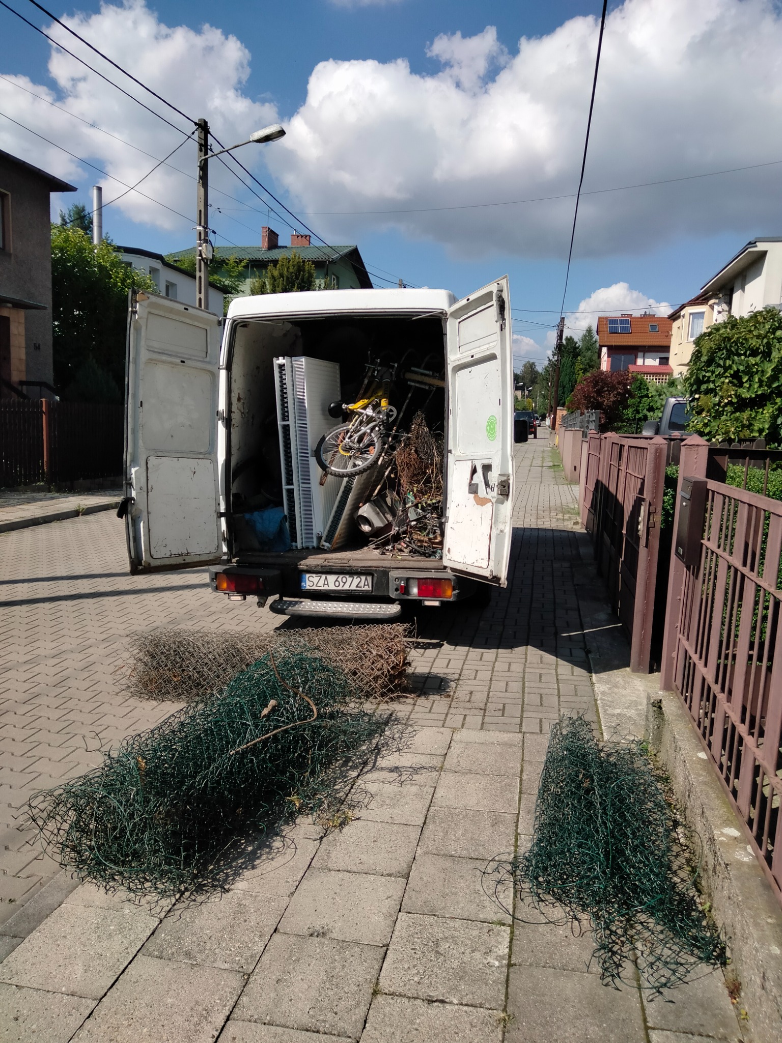 Otwarty furgon załadowany różnymi przedmiotami, w tym rowerem i grzejnikiem, zaparkowany na chodniku z rozwiniętą zieloną siatką ogrodzeniową na ziemi.