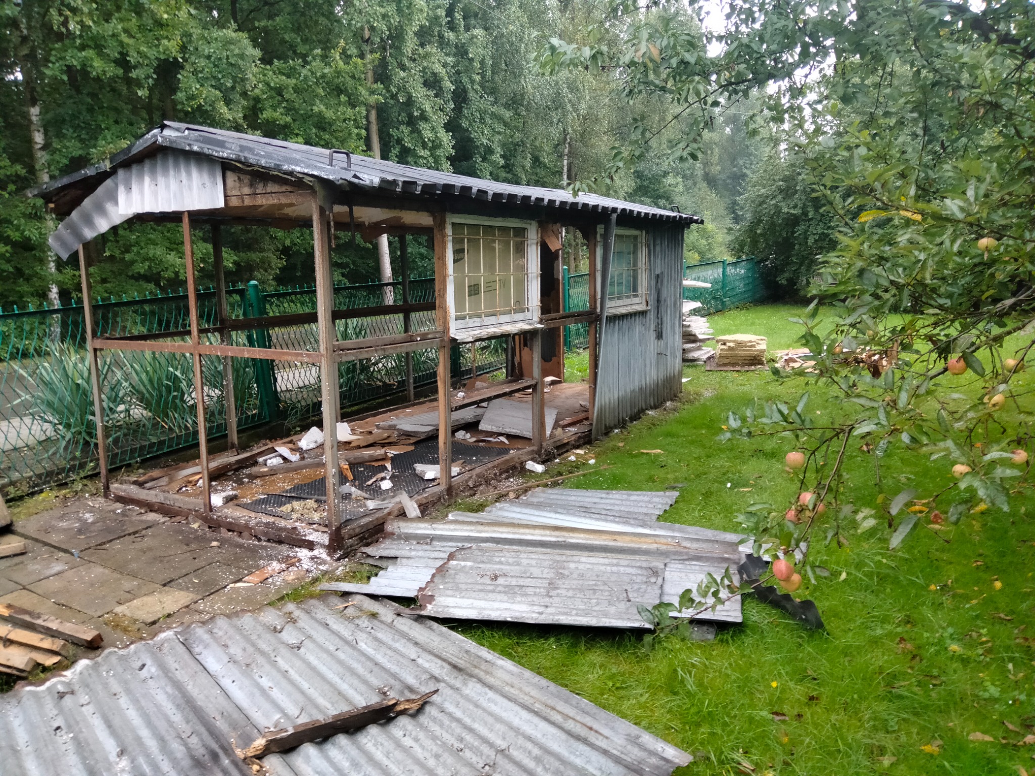 Rozbiórka drewnianej szopy z eternitowym dachem na zarośniętej działce, widoczne fragmenty konstrukcji, porozrzucane płyty i okna.