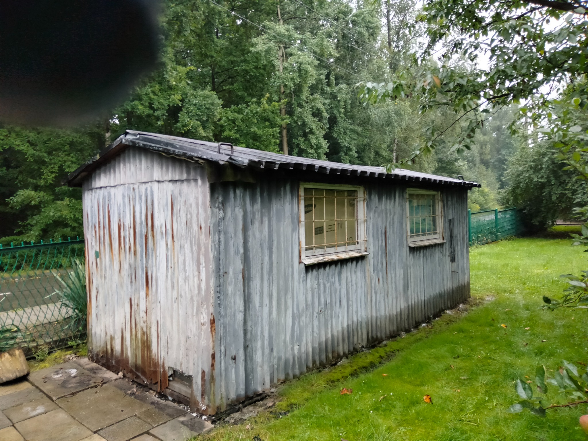 Zewnętrzny widok zniszczonego, blaszanego baraku z dwoma oknami z kratami, stojącego na skraju trawnika z widocznym fragmentem kortu tenisowego w tle.
