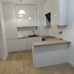 Nowoczesna, biała kuchnia z drewnianym blatem, czarnym zlewem i płytą indukcyjną, widoczne niedokończone prace wykończeniowe na ścianie.