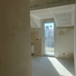 Wnętrze poddasza w trakcie prac wykończeniowych: ściany i sufit pokryte gładzią gipsową, widoczne okno dachowe z promieniem słońca na posadzce, grzejnik oraz przygotowane otwory na gniazdka...