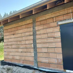 Wznoszenie ściany z pustaków ceramicznych na fundamencie z izolacją przeciwwilgociową, widoczny fragment dachu z rynną i czarne drzwi wejściowe.