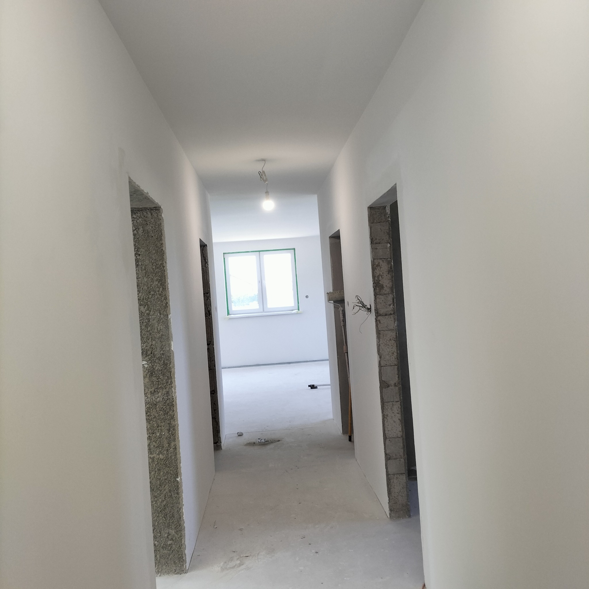 Korytarz w trakcie remontu: ściany otynkowane na biało, widoczne surowe ościeżnice drzwiowe, podłoga wylewka, w głębi okno z zieloną taśmą malarską.