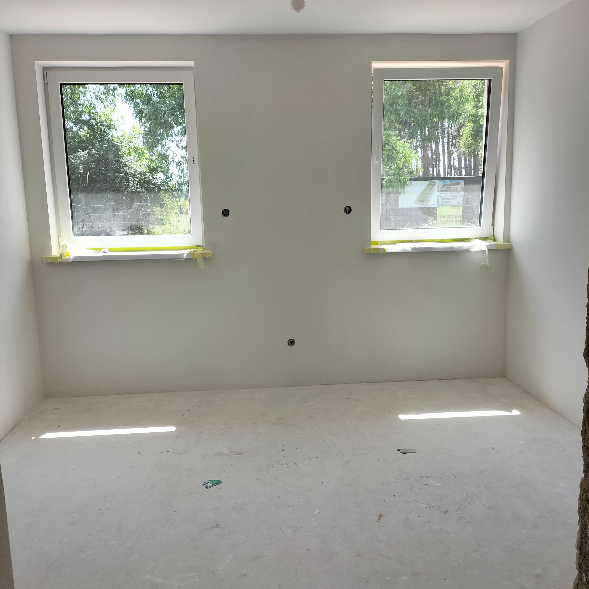 Wnętrze pokoju w stanie deweloperskim, z dwoma oknami zabezpieczonymi taśmą malarską, gładkie, białe ściany z widocznymi otworami pod instalacje elektryczne, surowa podłoga.