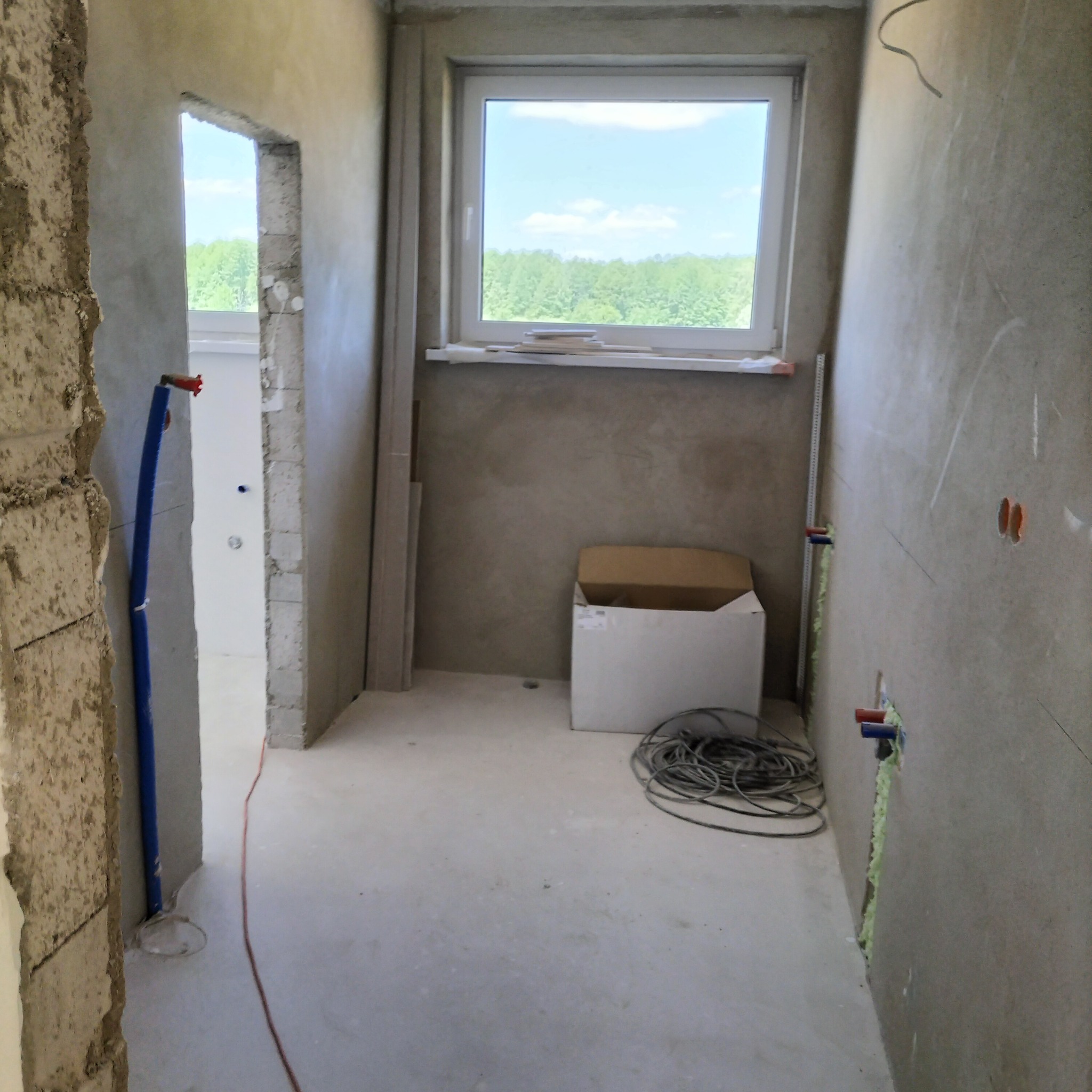 Wnętrze łazienki w trakcie remontu: surowe ściany, widoczne instalacje wodne, okno z widokiem na zieleń, karton i zwój kabli na podłodze.
