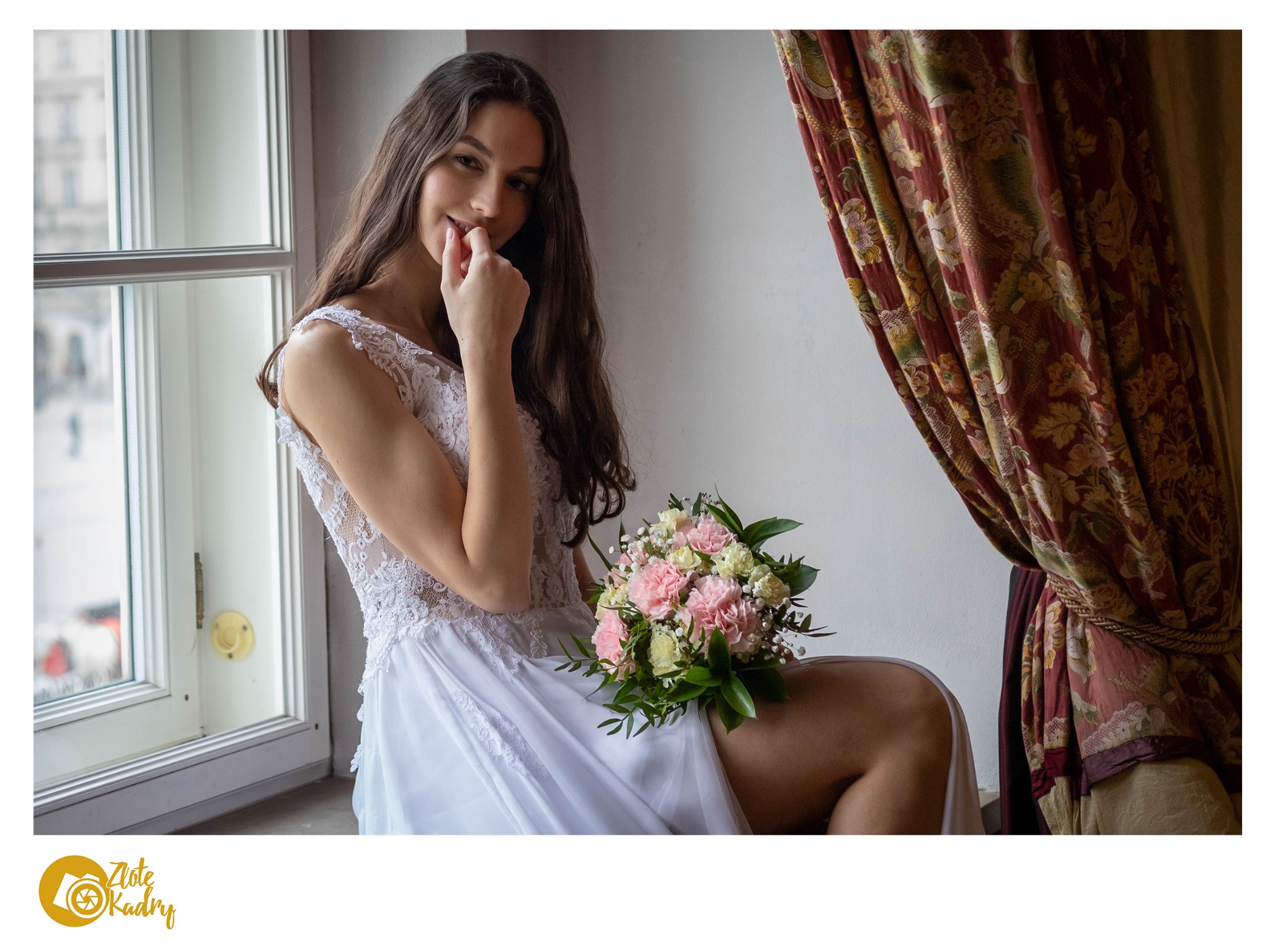 Uśmiechnięta panna młoda w koronkowej sukni siedzi w oknie, trzyma bukiet różowych i białych goździków, za nią dekoracyjne zasłony.