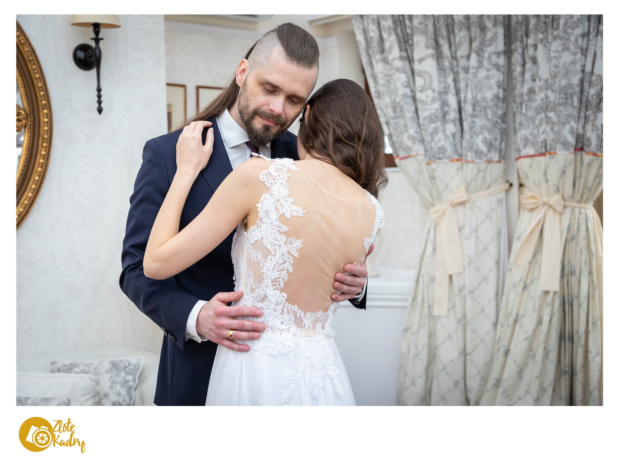 Para młoda w objęciach: panna młoda w sukni z koronkowym tyłem, pan młody w granatowym garniturze z oryginalną fryzurą, w tle wzorzyste zasłony i złote lustro.