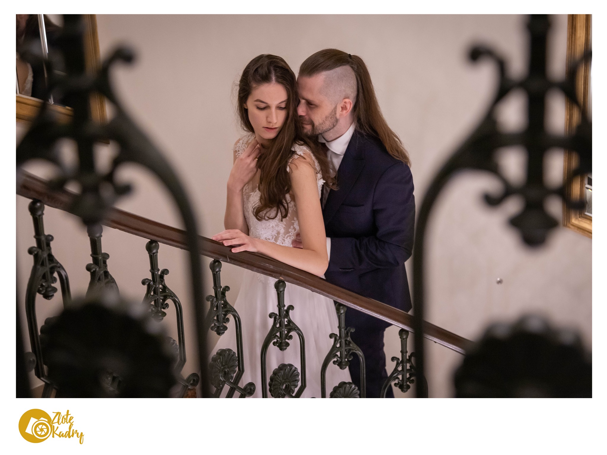 Para młoda w eleganckich strojach pozuje na schodach z ozdobną balustradą, panna młoda w sukni z koronkowymi detalami, pan młody w granatowym garniturze, subtelne światło podkreśla intymną atmosferę.