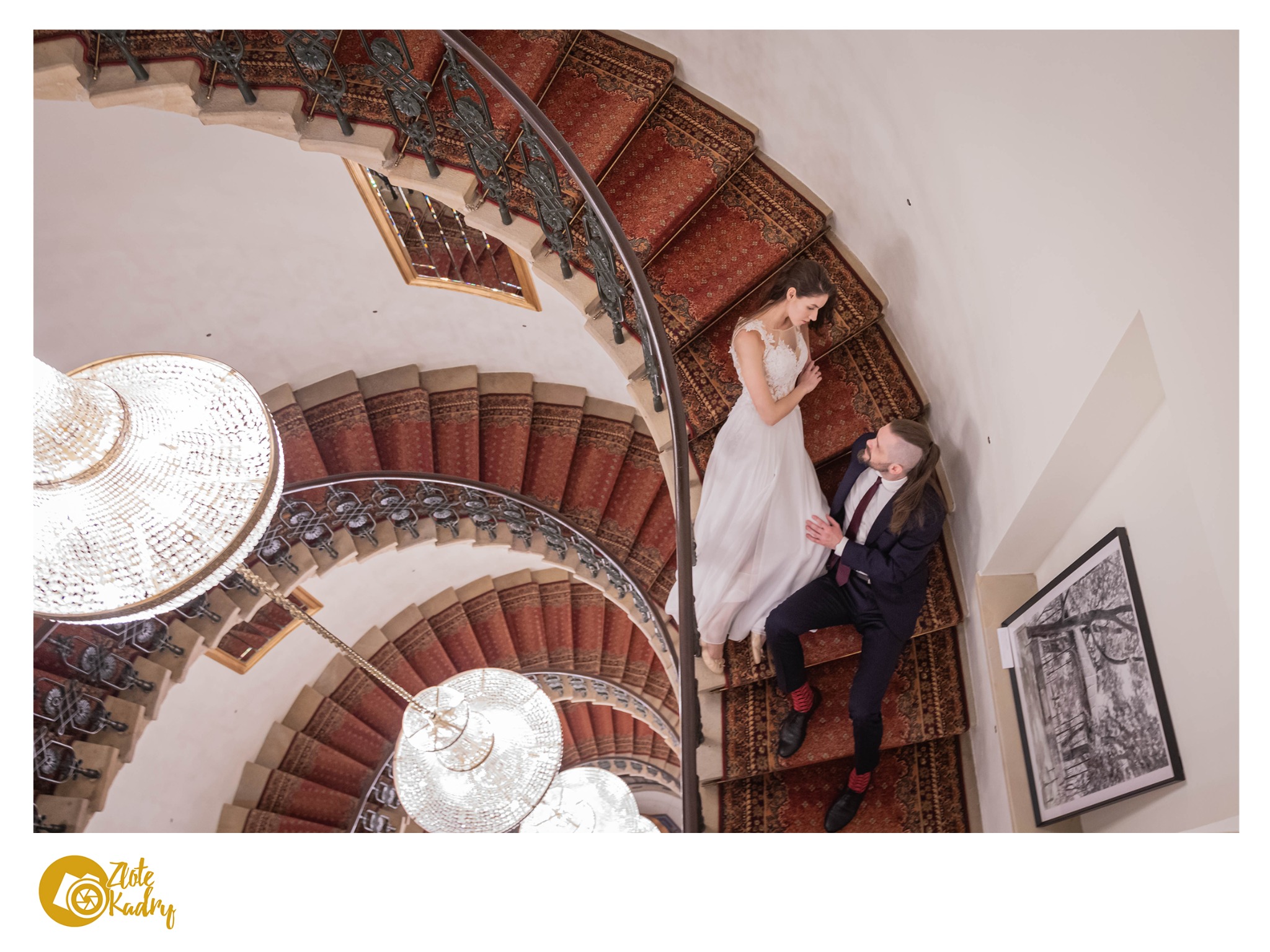 Para młoda siedzi na spiralnych schodach z czerwonym dywanem, widok z góry, kryształowe żyrandole w tle, na ścianie czarno-biała grafika w ramie.