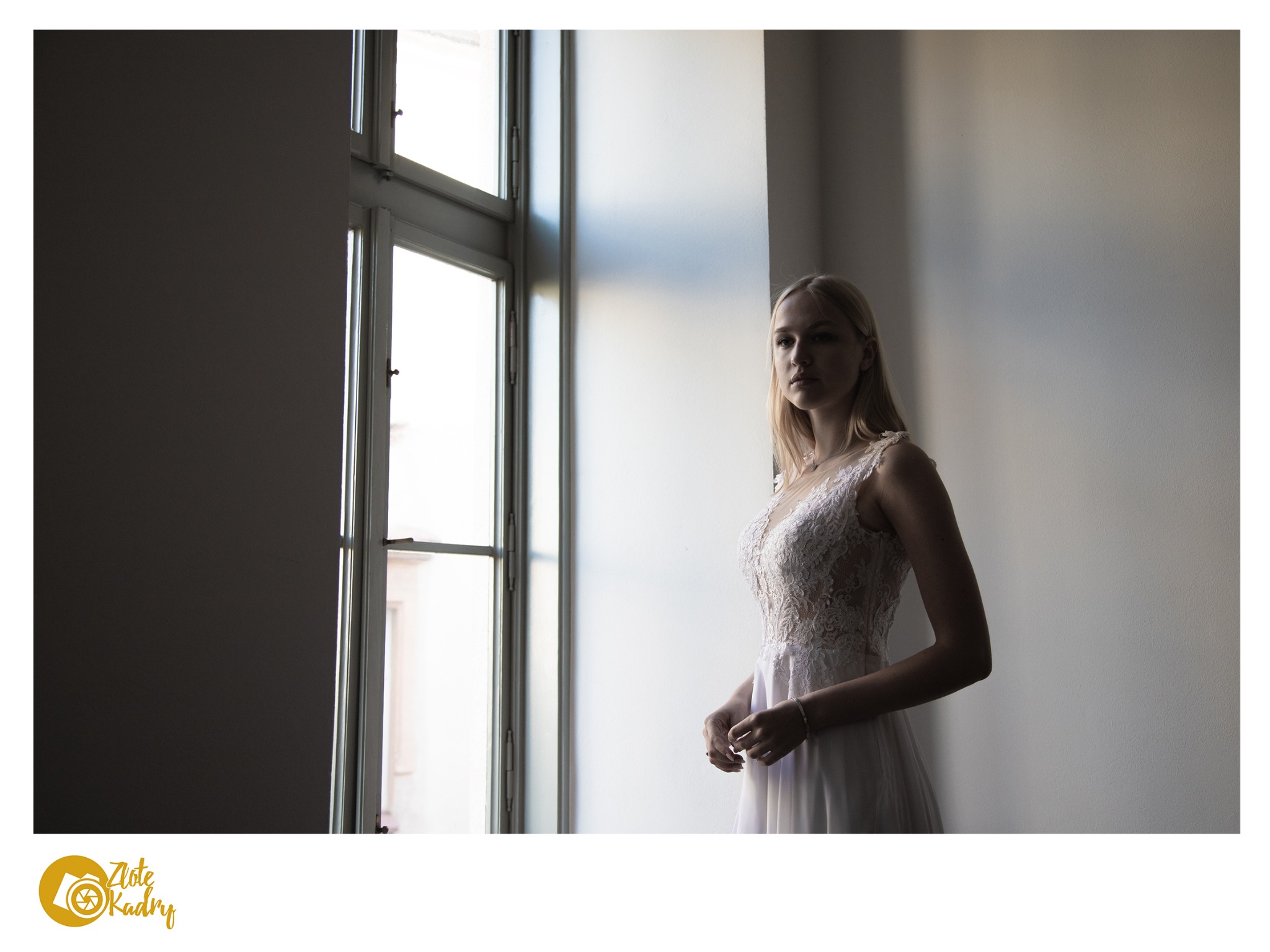 Panna młoda w koronkowej sukni, stojąca przy oknie z delikatnym światłem, subtelny portret w atelier.