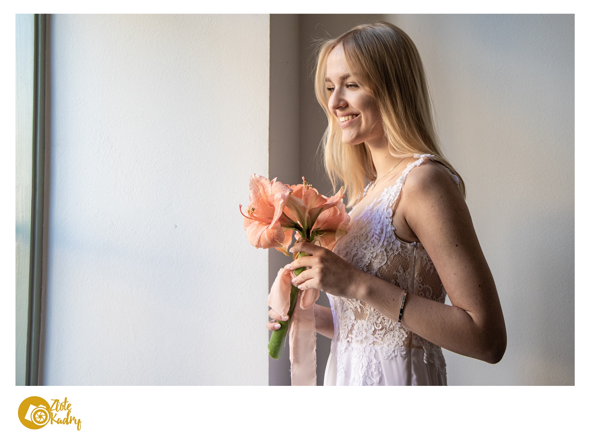 Uśmiechnięta panna młoda w koronkowej sukni, trzymająca bukiet amarylisów z delikatną wstążką, spogląda przez okno w słoneczny dzień.