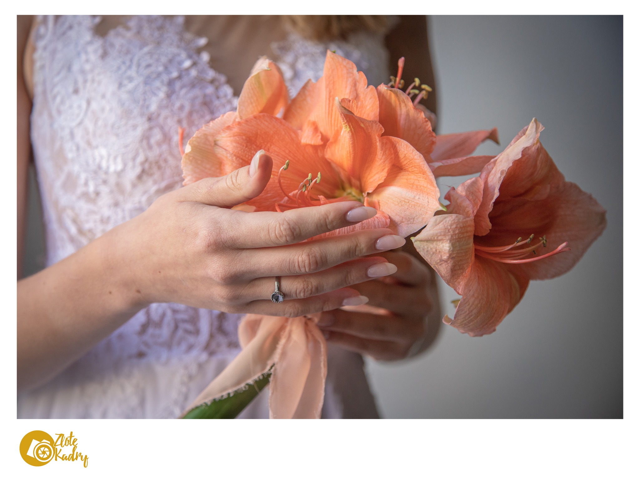 Panna młoda w koronkowej sukni trzyma bukiet amarylisów w odcieniach brzoskwini, eksponowany pierścionek zaręczynowy na palcu.