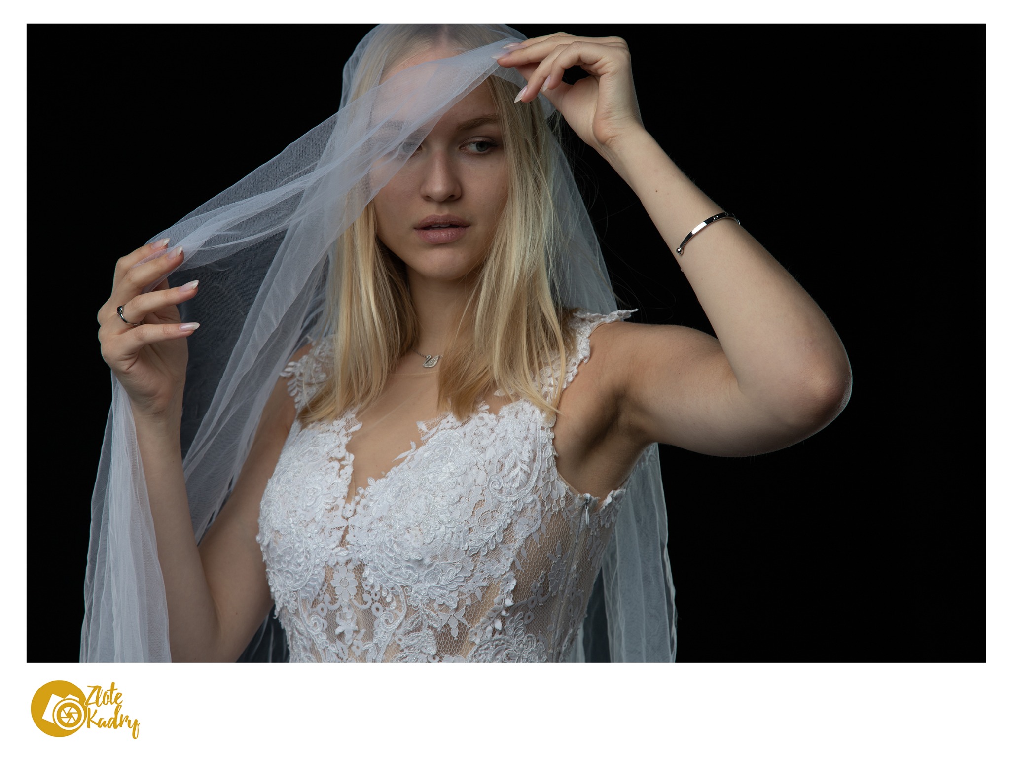 Panna młoda w koronkowej sukni i welonie, z subtelną biżuterią na szyi i nadgarstku, pozuje na czarnym tle.