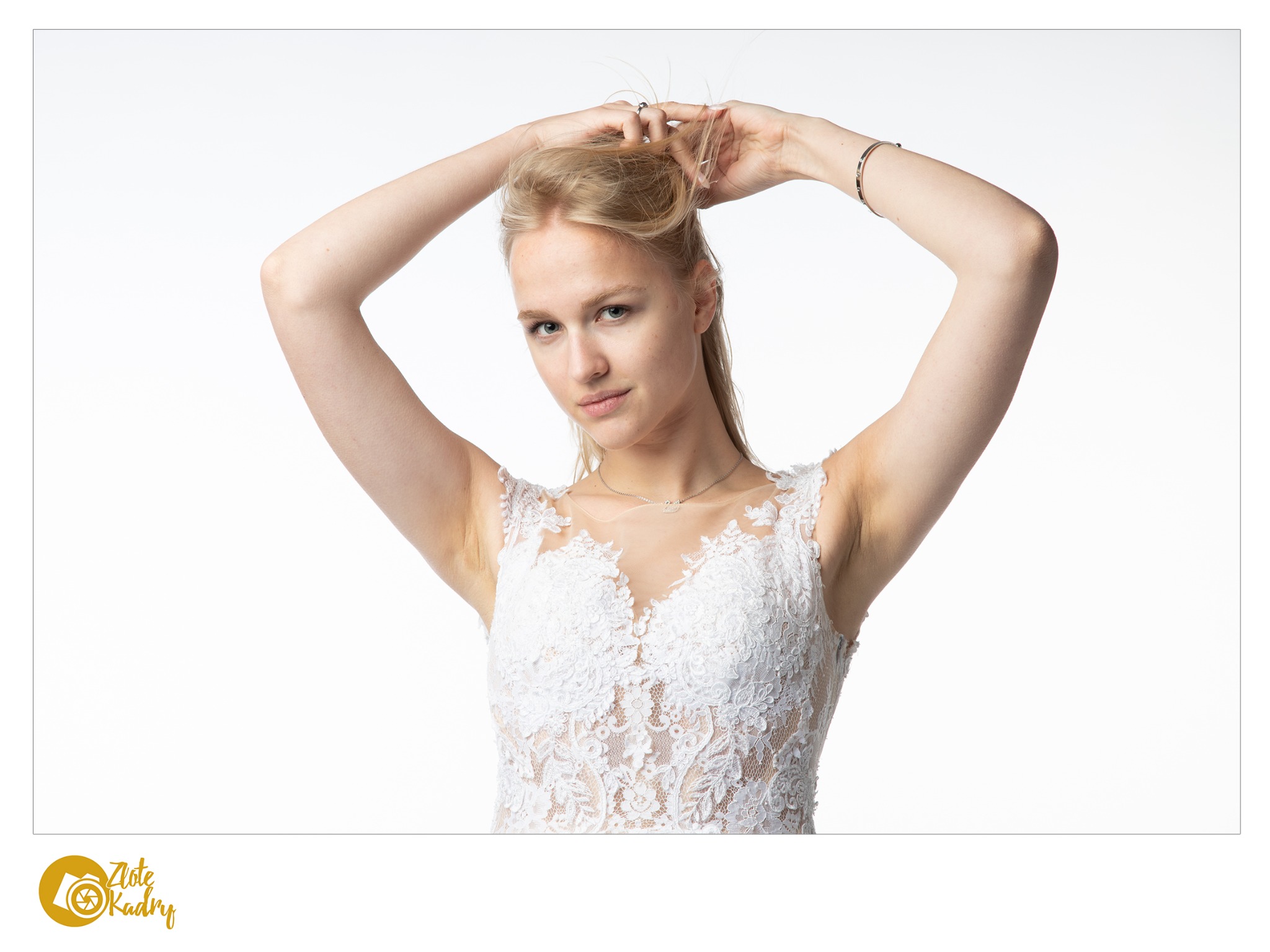 Portret kobiety w koronkowej sukni, z rękami uniesionymi do góry, poprawiającej włosy na białym tle.