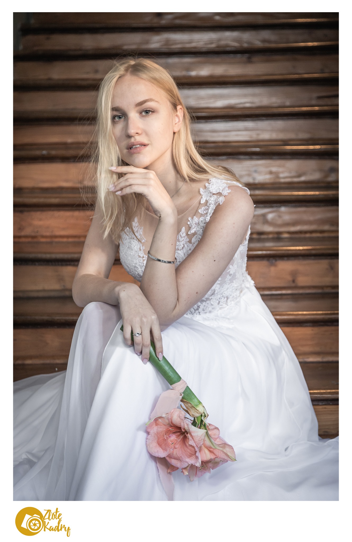 Panna młoda w białej sukni z koronkową górą, siedząca na drewnianych schodach, trzymająca bukiet amarylisów z różową wstążką.