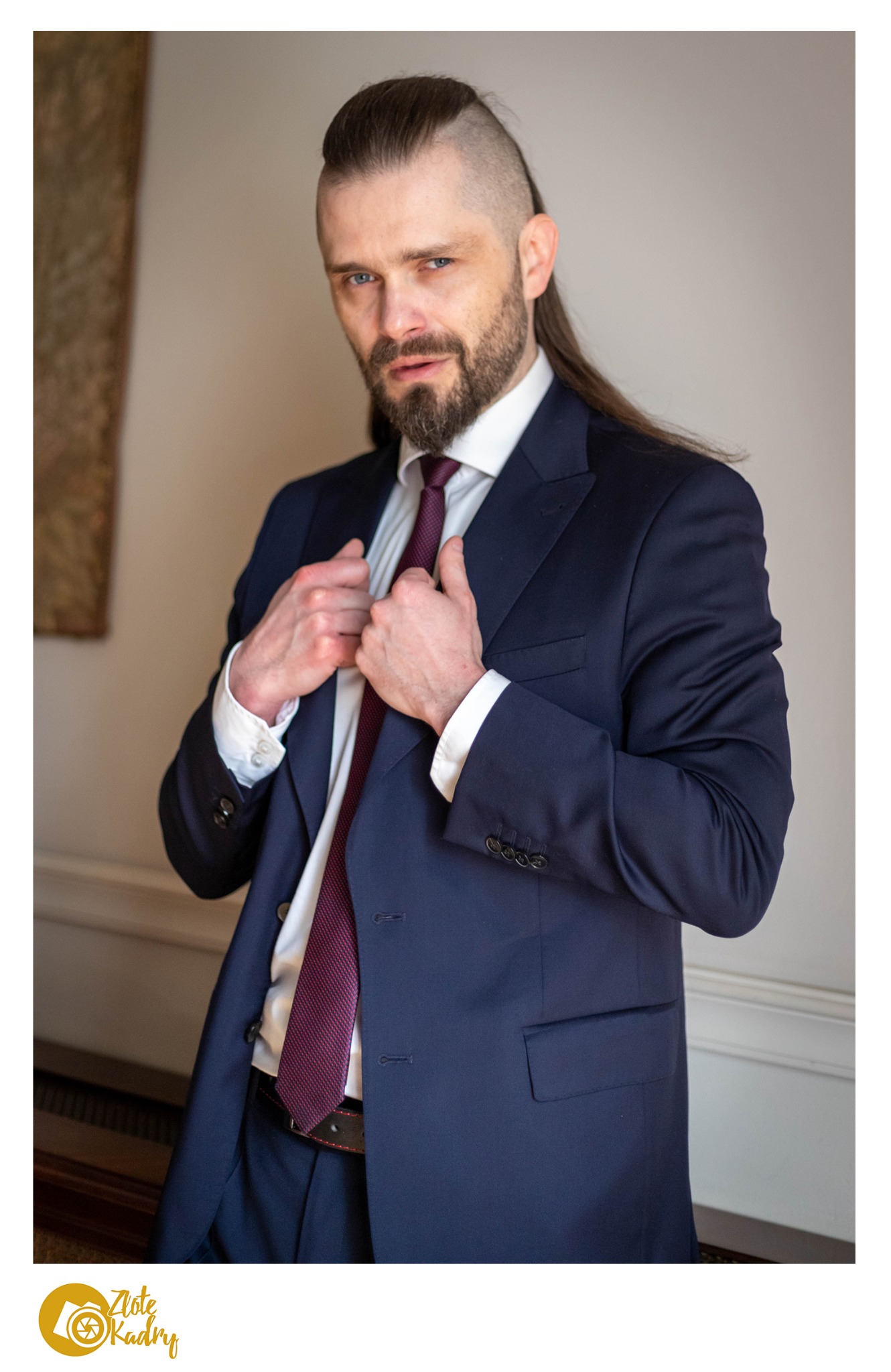 Mężczyzna w granatowym garniturze i bordowym krawacie, z charakterystyczną fryzurą, pozuje do portretu w neutralnym wnętrzu.