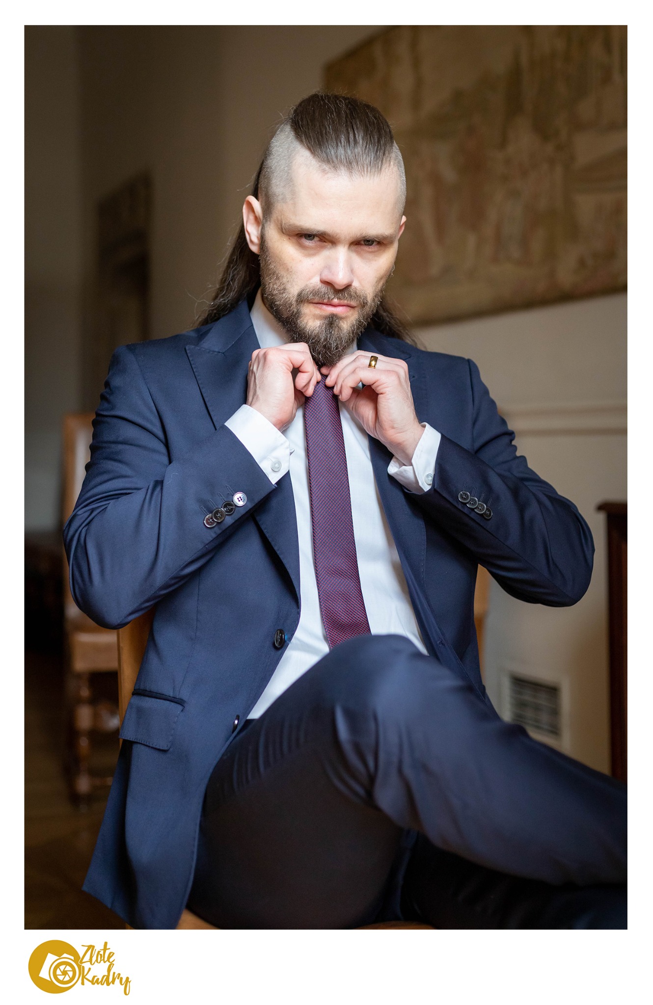 Portret mężczyzny z brodą i długimi włosami, ubrany w granatowy garnitur, poprawiającego bordowy krawat, siedzącego na drewnianym krześle w pomieszczeniu z obrazem w tle.