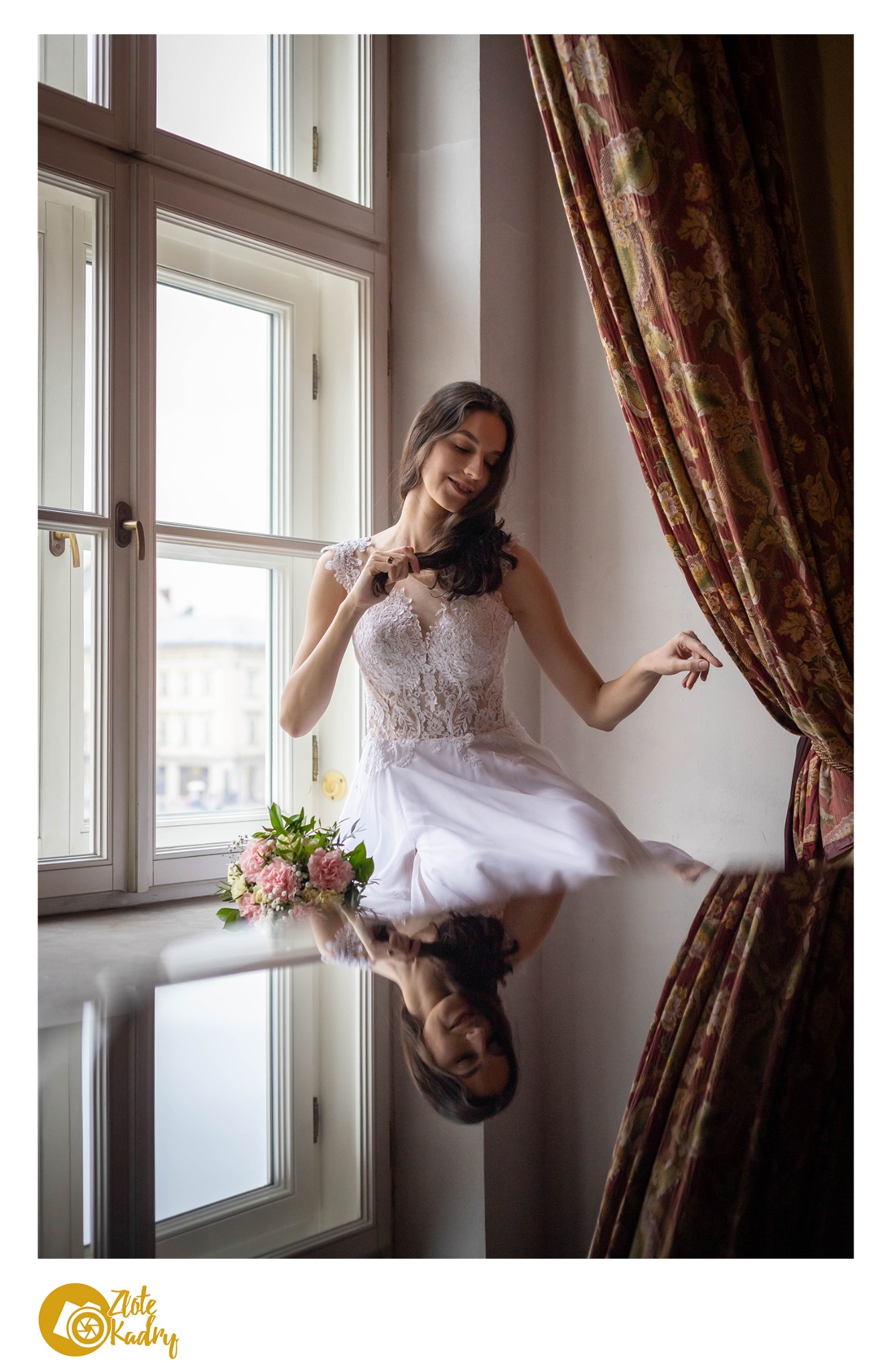 Panna młoda w koronkowej sukni pozuje przy oknie z bukietem, odbicie w błyszczącej powierzchni, dekoracyjne zasłony w tle.