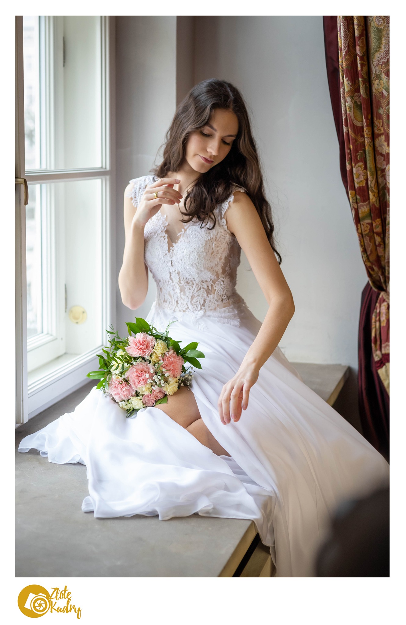 Panna młoda w koronkowej sukni siedzi na parapecie okna, trzymając bukiet różowych i białych goździków, z dekoracyjną zasłoną w tle.