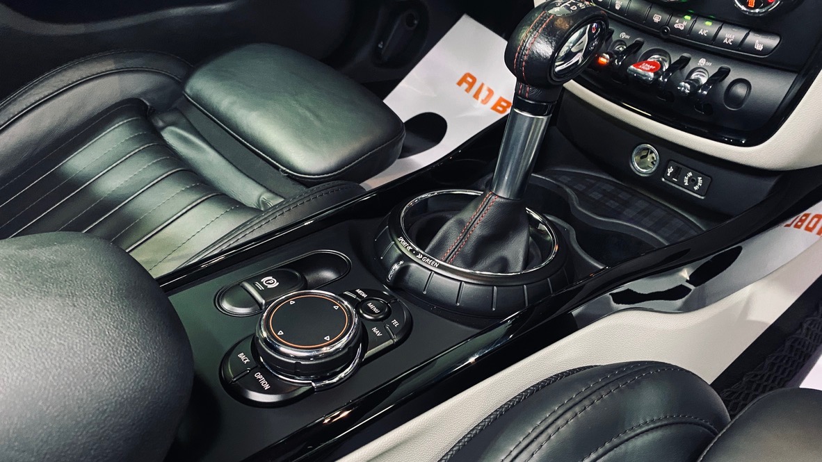 Wnętrze samochodu Mini Cooper z czarną skórzaną tapicerką, widoczny kokpit, dźwignia zmiany biegów z czerwoną nicią i panele sterowania, uchwycone w jasnym świetle.