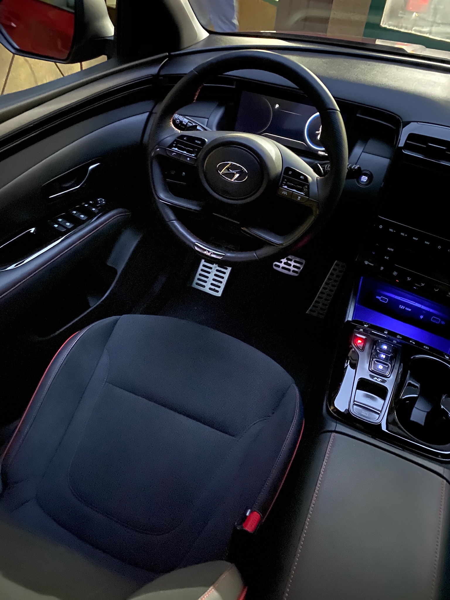 Wnętrze samochodu Hyundai Tucson N Line z czarną tapicerką i czerwonymi przeszyciami, widok na kierownicę, deskę rozdzielczą z cyfrowym wyświetlaczem i metalowe nakładki na pedały.