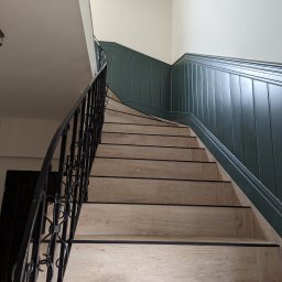 Lech-Bud - Klatka schodowa z drewnopodobnymi stopniami i czarną, kutą balustradą, widok z dołu ku górze, ściany wykończone boazerią w odcieniach zieleni i bieli.