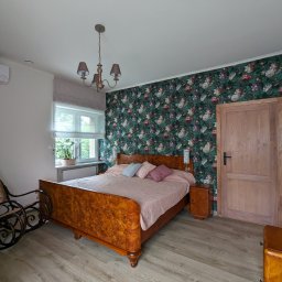 Lech-Bud - Sypialnia z tapetą w kwiaty, drewnianym łóżkiem z różową narzutą i krzesłem bujanym, oświetlona przez żyrandol z beżowymi abażurami i światło dzienne wpadające przez okno z roletą rzymską.