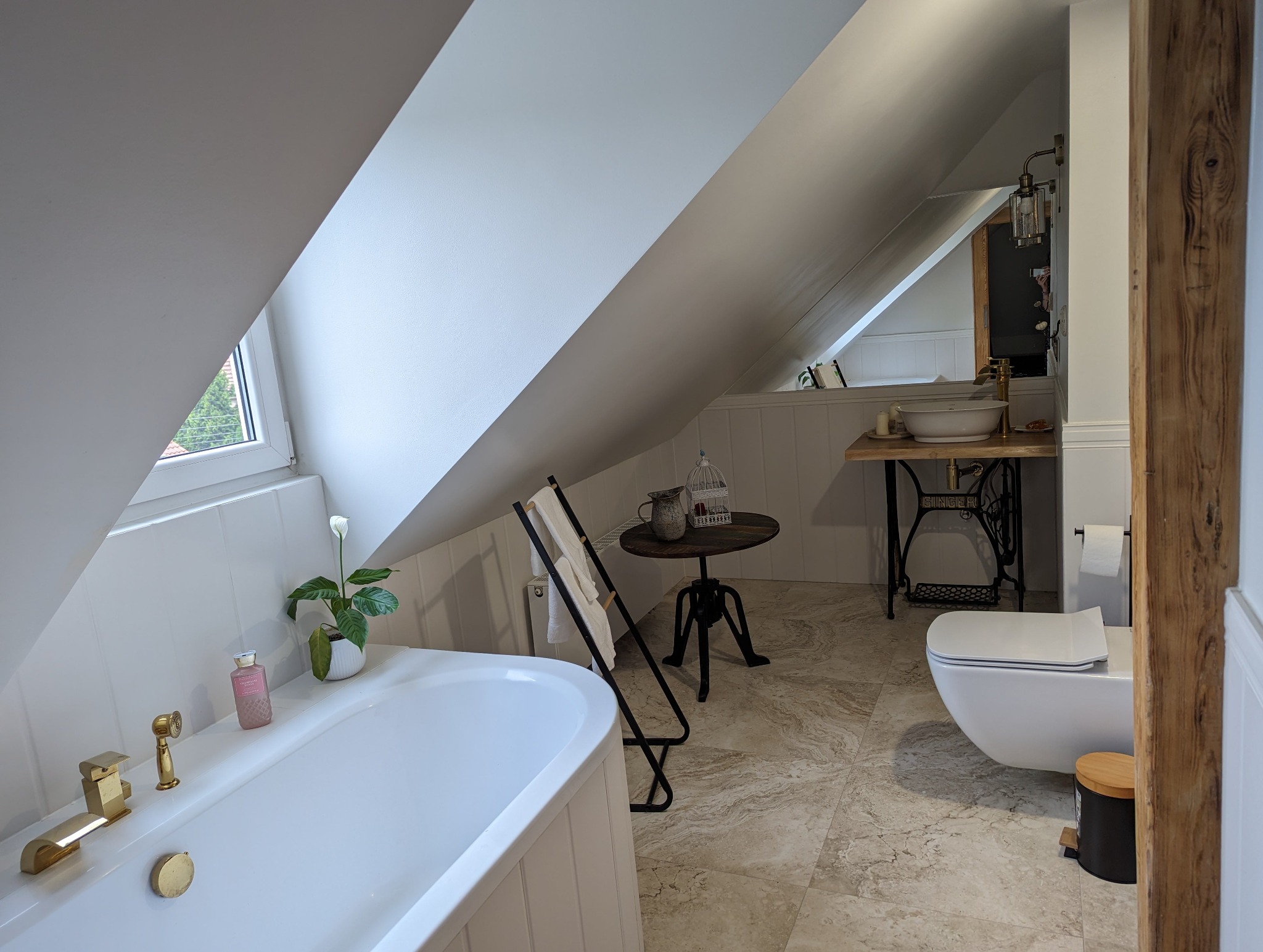 Jasna łazienka na poddaszu ze skośnym sufitem, z wanną, toaletą, umywalką na postumencie z maszyny do szycia Singer, stolikiem z klatką dla ptaków i ręcznikami na wieszaku.