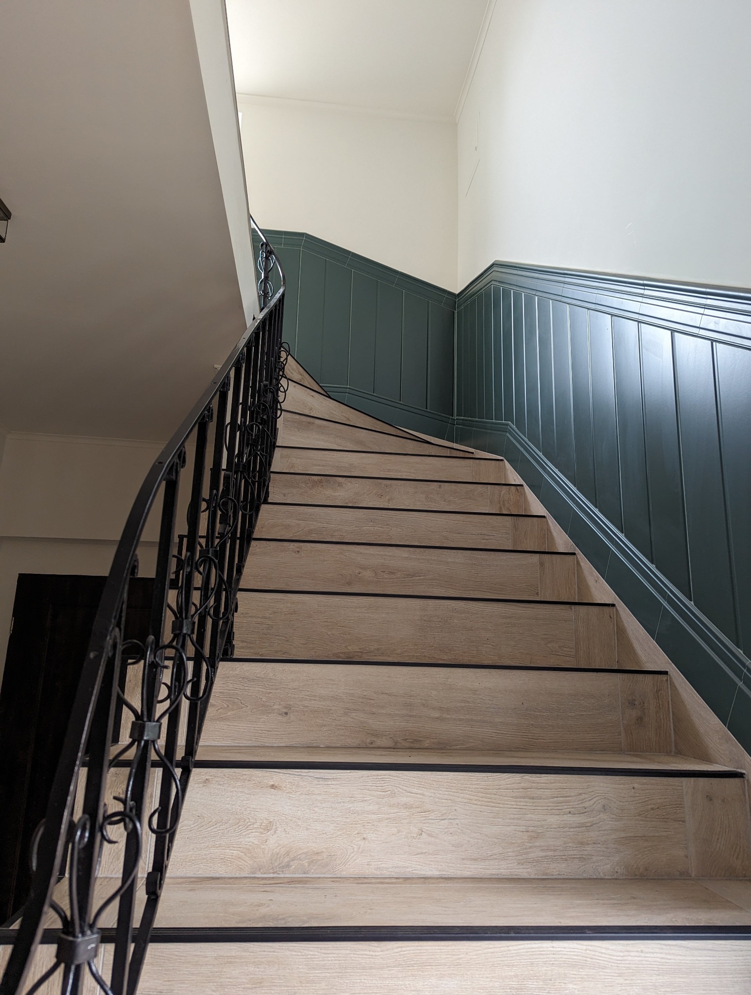 Klatka schodowa z drewnopodobnymi stopniami i czarną, kutą balustradą, widok z dołu ku górze, ściany wykończone boazerią w odcieniach zieleni i bieli.