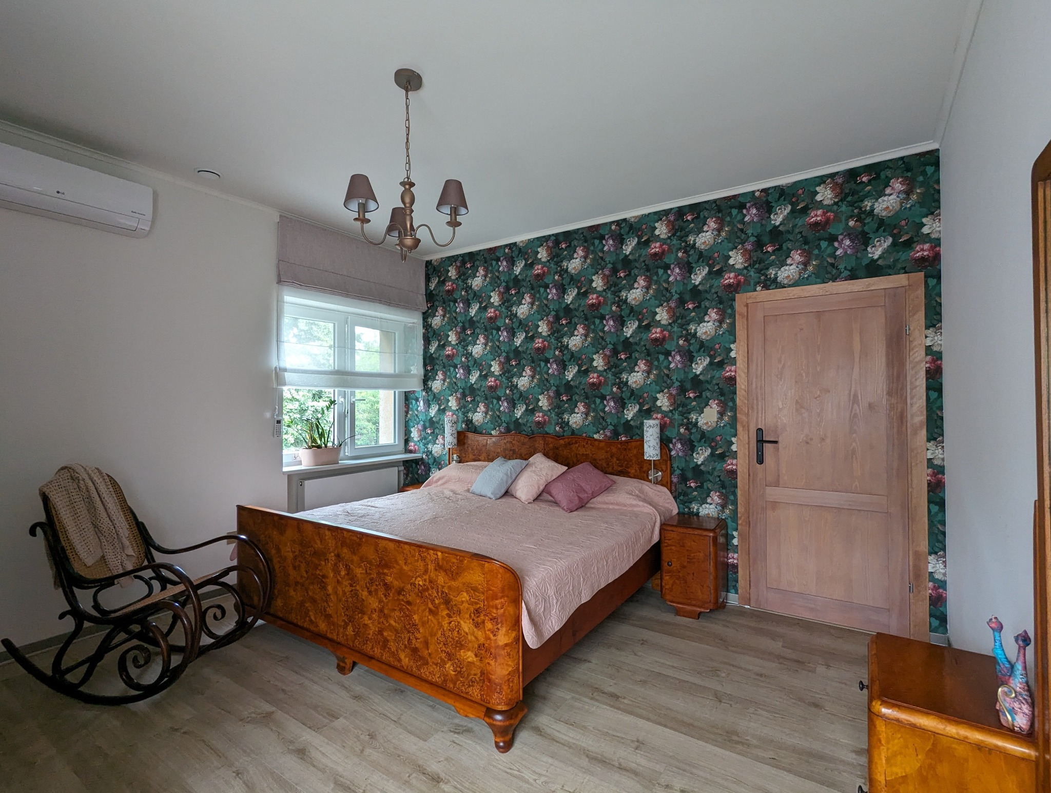 Sypialnia z tapetą w kwiaty, drewnianym łóżkiem z różową narzutą i krzesłem bujanym, oświetlona przez żyrandol z beżowymi abażurami i światło dzienne wpadające przez okno z roletą rzymską.