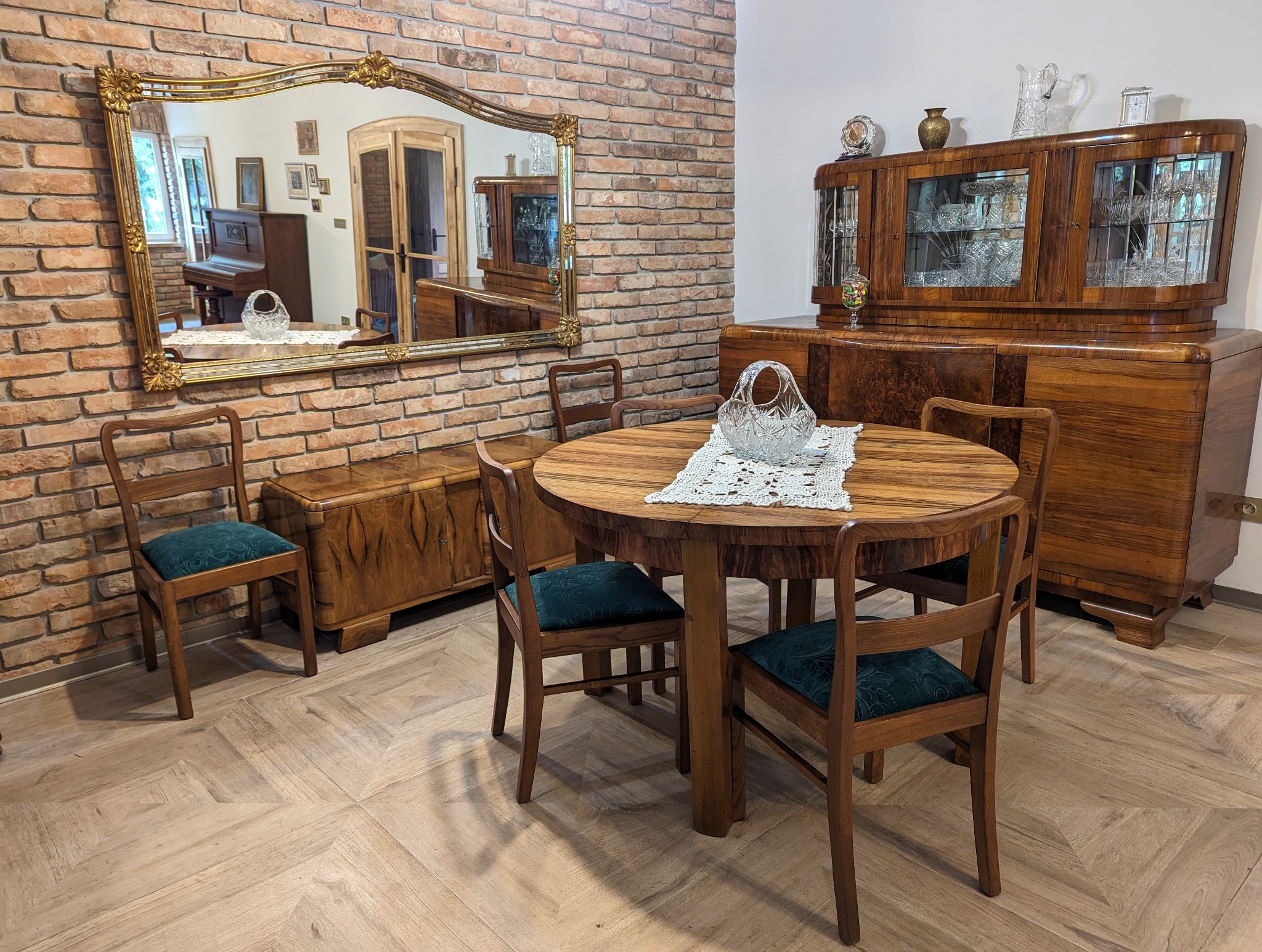 Salon z odrestaurowanymi meblami w stylu art deco: okrągły stół z kryształowym koszyczkiem na serwecie, krzesła z zielonym obiciem, kredens i komoda z fornirem oraz lustro w złotej ramie...