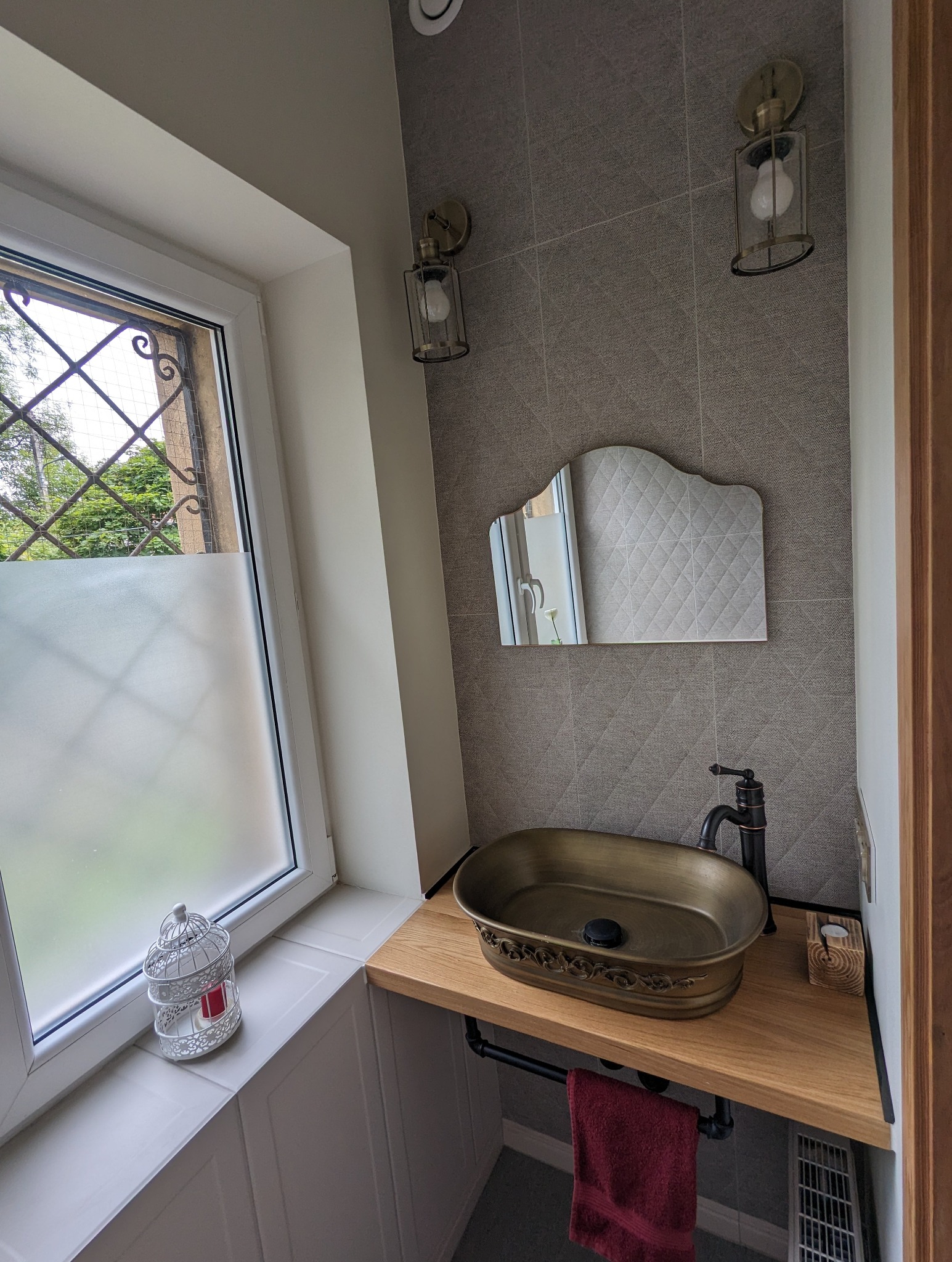 Aranżacja małej łazienki z umywalką w stylu retro, armaturą w kolorze starego brązu, lustrem w ozdobnej ramie i lampami ściennymi w stylu industrialnym.