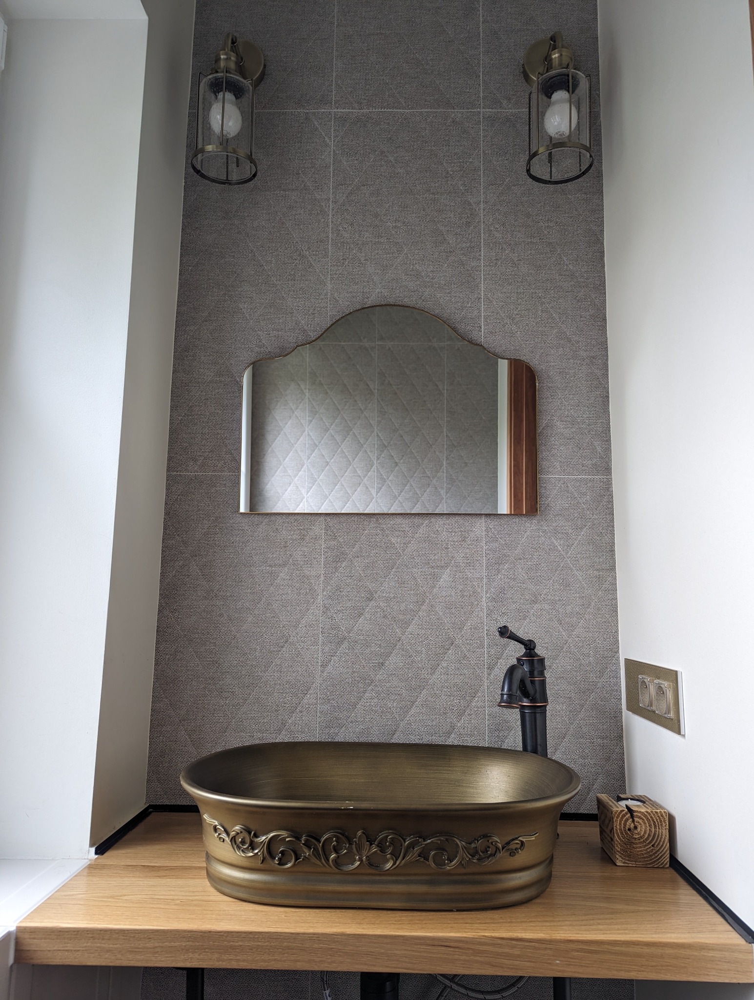 Wykończona łazienka z umywalką w stylu retro, armaturą w kolorze starego złota i lustrem o nietypowym kształcie na tle szarej ściany z płytek.