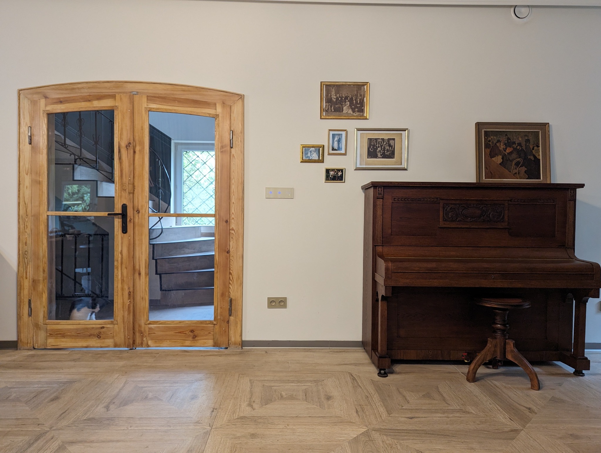 Drewniane dwuskrzydłowe drzwi z szybami, przez które widać spiralne schody i siedzącego kota, na ścianie galeria rodzinnych zdjęć i pianino ze stołkiem, na podłodze parkiet w geometryczny wzór.